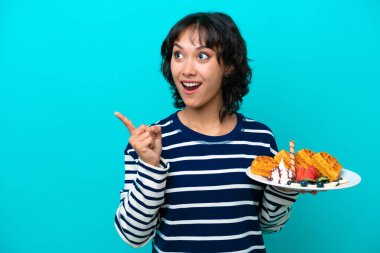 Arjantinli genç bir kadın mavi arka planda izole edilmiş waffleları tutarken bir parmağını kaldırarak çözümü anlamaya çalışıyor.