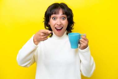 Arjantinli genç bir kadın elinde kahve fincanı tutarken sarı arka planda izole edilmiş şaşırmış ve ön tarafı işaret ediyor.