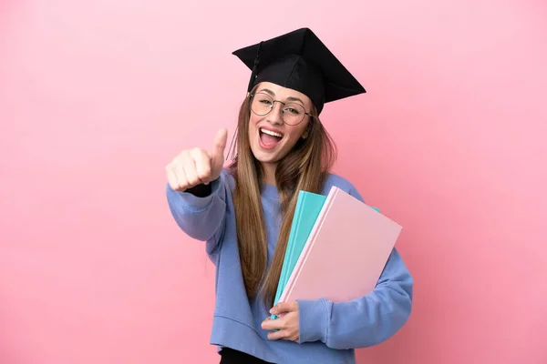 若いです学生女性身に着けていますA大学院生帽子絶縁上のピンクの背景とともに親指アップ何か良いことが起こっているので — ストック写真
