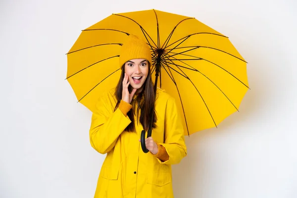 驚きとショックを受けた表情で白い背景に隔離されたレインプルーフコートと傘を持つ若いブラジル人女性 — ストック写真