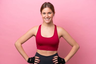 Pembe arka planda izole edilmiş genç beyaz kadın kolları kalçasında ve gülümsüyor.