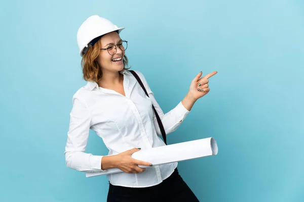 Young architect Georgian woman with helmet and holding blueprints over isolated background pointing finger to the side and presenting a product