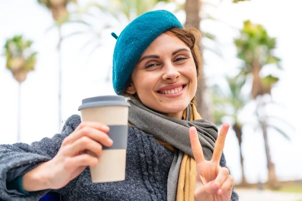 布鲁内特的女人微笑着在户外拿起咖啡 并展示胜利的标志 — 图库照片
