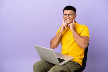 Genç adam dizüstü bilgisayarlı bir sandalyede oturmuş mutlu ve hoş bir ifadeyle gülümsüyor.