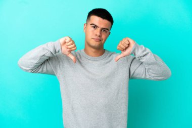 Soyutlanmış mavi arka planda genç yakışıklı adam baş parmağını iki eliyle aşağı gösteriyor.