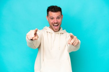 Mavi arka planda izole edilmiş beyaz, yakışıklı genç bir adam şaşırmış ve ön tarafı işaret ediyor.