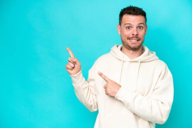 Kafkasyalı yakışıklı adam mavi arka planda izole edilmiş, korkmuş ve kenarı işaret ediyor.