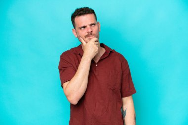 Kafkasyalı yakışıklı bir adam mavi arka planda izole edilmiş. Şüpheleri var ve kafası karışmış bir yüz ifadesi var.
