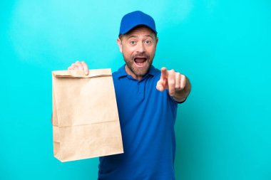 Orta yaşlı bir adam mavi arka planda izole edilmiş bir paket yiyecek alıyor. Şaşırmış ve ön tarafı işaret ediyor.