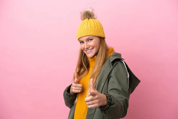 年轻的金发女人穿着冬季夹克 背景是粉色的 她指着前面笑着 — 图库照片