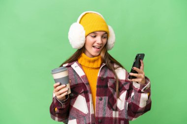 Kafkasyalı genç bir kız izole edilmiş arka planda kışlık kuklalar takıyor. Elinde kahve ve cep telefonu var.