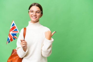 Kafkasyalı genç bir kız izole edilmiş arka planda Birleşik Krallık bayrağı tutuyor ve bir ürün sunmak için kenarı işaret ediyor.