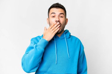 İzole edilmiş beyaz arka planda genç, yakışıklı beyaz bir adam esniyor ve ağzını eliyle kapatıyor.