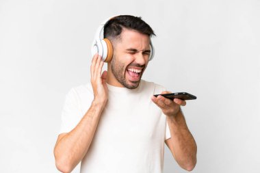 İzole edilmiş beyaz arka planda genç, yakışıklı beyaz bir adam cep telefonuyla müzik dinliyor ve şarkı söylüyor.
