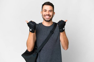 Spor çantalı beyaz tenli genç beyaz bir adam. Başparmakları havada ve gülümsüyor.