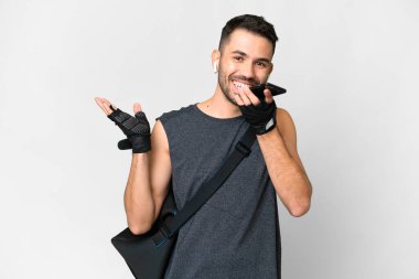 İzole edilmiş beyaz arka planda spor çantalı beyaz bir genç adam cep telefonuyla sohbet ediyor.