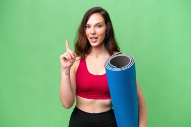 Genç sporcu kadın yoga derslerine giderken izole bir arka plan üzerinde minder tutuyor ve parmağıyla işaret eden bir fikir düşünüyor.