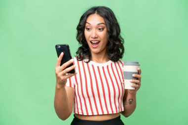 Arjantinli genç bir kadın izole edilmiş arka planda kahve ve bir cep telefonu tutuyor.