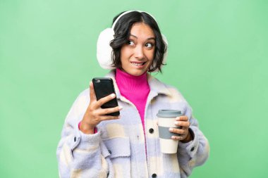 Arjantinli genç bir kadın izole edilmiş arka planda kışlık kuklalar takıyor. Elinde kahve ve bir şeyler düşünürken cep telefonu var.