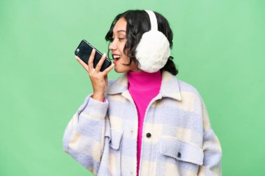 Arjantinli genç bir kadın izole edilmiş arka planda kışlık manşetler takıyor ve cep telefonuyla sohbet ediyor.