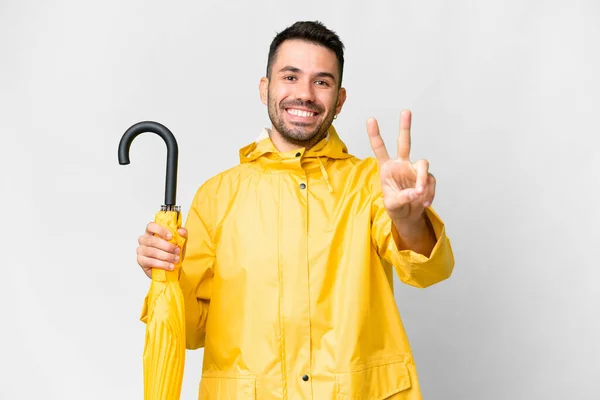 年轻的高加索人 身穿防雨外套 披着雨伞 面带微笑 露出胜利的迹象 — 图库照片