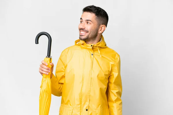 年轻的高加索人 身穿防雨外套 带着雨伞 披着孤零零的白色背景 朝旁边看去 微笑着 — 图库照片