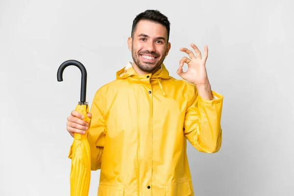 年轻的高加索人 身穿防雨外套 带着雨伞 盖在孤独的白色背景上 用手指显示出可控的标志 — 图库照片