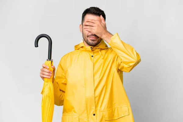 手で目をカバー隔離された白い背景の上にレインプルーフコートと傘を持つ若い白人男性 何か見たくない — ストック写真
