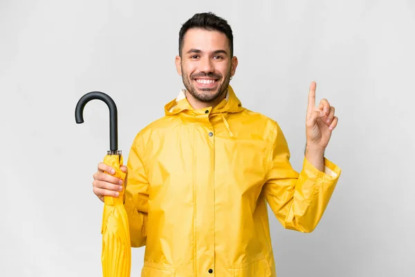 年轻的高加索人 身穿防雨外套 带着雨伞 盖在孤立的白色背景上 展示并举起一个手指 表示他是最好的象征 — 图库照片