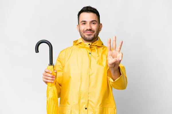 年轻的高加索人 穿着防雨外套 带着雨伞 高高兴兴地盖在孤立的白色背景上 用手指数着三个 — 图库照片