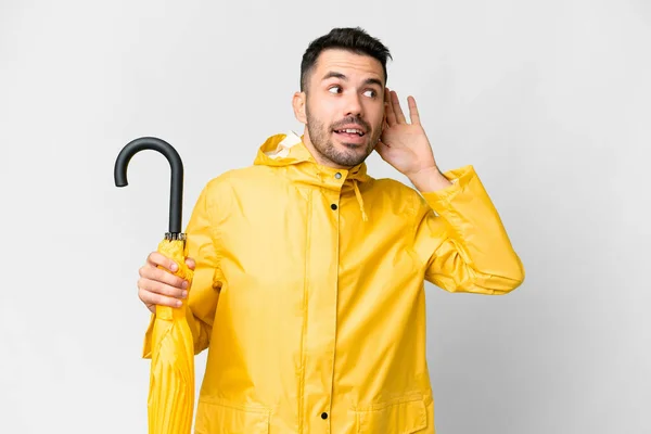 耳に手を置くことによって何かを聞く隔離された白い背景の上にレインプルーフコートと傘を持つ若い白人男性 — ストック写真