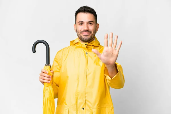 年轻的高加索人 身穿防雨外套 带着雨伞 盖在孤立的白色背景上 手里拿着五根手指 — 图库照片