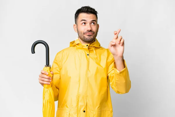 年轻的高加索人 身穿防雨外套 带着雨伞 盖在孤立的白色背景上 手指头交叉 祝福好运 — 图库照片