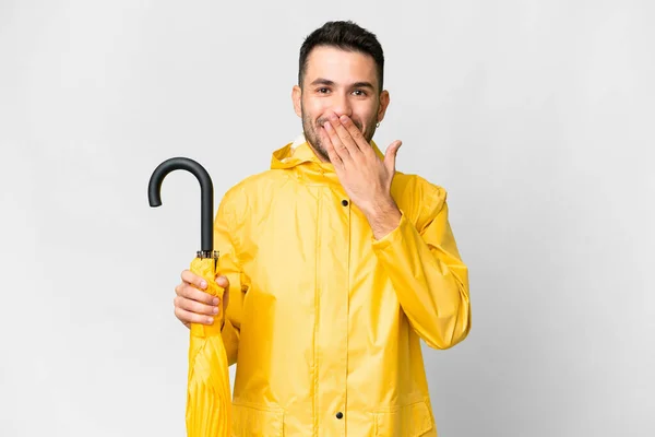 年轻的高加索人 身穿防雨外套 披着雨伞 披着孤零零的白色背景 幸福地微笑着 用手捂着嘴 — 图库照片