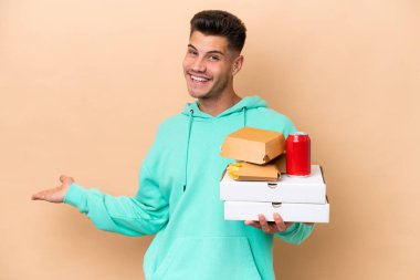 Kafkasyalı genç adam bej arka planda izole edilmiş fast food tutuyor. Davet etmek için ellerini yana açıyor.