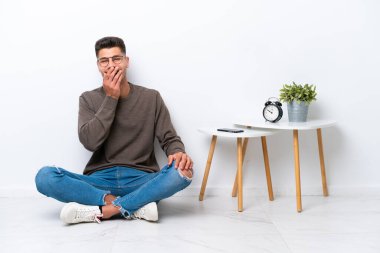 Beyaz arka planda yalnız başına oturan beyaz tenli genç adam mutlu ve gülümsüyor.