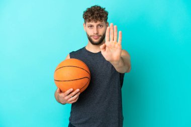 Basketbol oynayan yakışıklı genç adam mavi arka planda izole edilmiş bir şekilde durup jest yapıyor.