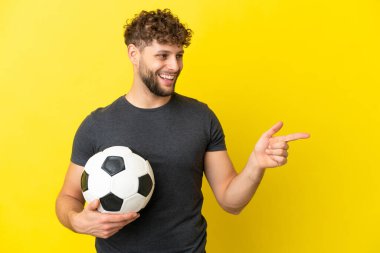 Yakışıklı genç futbolcu sarı arka planda izole edilmiş, parmağını yana doğru gösteren ve bir ürün sunan.