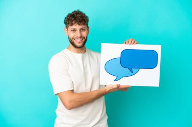 Genç, beyaz, mavi arka planda izole edilmiş genç bir adam elinde mutlu ifadeli bir konuşma baloncuğu ikonu olan bir levha tutuyor.