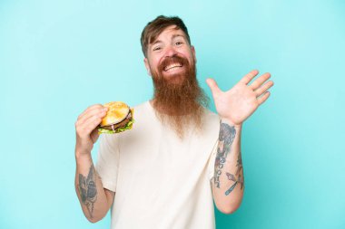 Kızıl saçlı, uzun sakallı bir adam elinde hamburgerle mavi arka planda izole edilmiş mutlu bir ifadeyle selam veriyor.