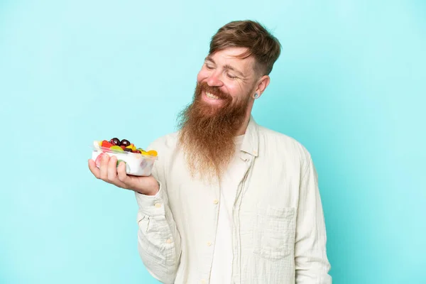 長い髭を生やした赤毛の男は幸せな表情で青い背景に隔離された果物のボウルを持っています — ストック写真