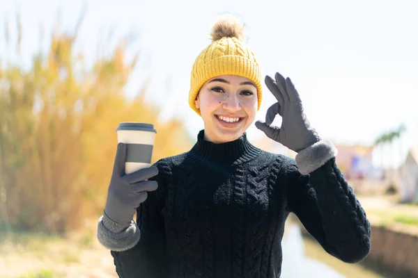 Young moroccan girl wearing winter muffs while holding a coffee at outdoors showing ok sign with fingers