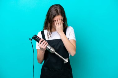 Beyaz tenli genç aşçı kadın el blenderi kullanıyor. Mavi arka planda izole edilmiş, yorgun ve hasta bir yüz ifadesi var.