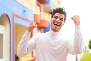 Genç beyaz adam dışarıda gözlüklü ve zaferi kutluyor.