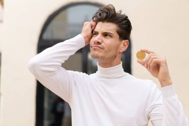 Kafkasyalı genç bir adam Bitcoin 'i açık havada tutuyor şüpheleri var ve kafası karışmış bir yüz ifadesi var.