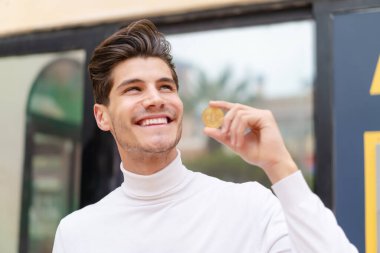 Açık havada elinde Bitcoin olan genç beyaz bir adam yukarı bakıyor.