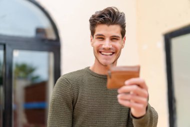 Genç beyaz bir adam açık havada mutlu bir ifadeyle bir cüzdan tutuyor.