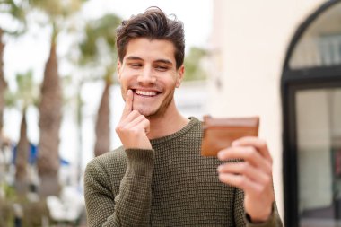 Açık havada genç beyaz bir adam elinde mutlu bir ifadeyle bir cüzdan tutuyor.