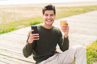 Açık havada cep telefonu kullanan ve elinde mutlu bir ifadeyle kahve tutan genç beyaz bir adam.