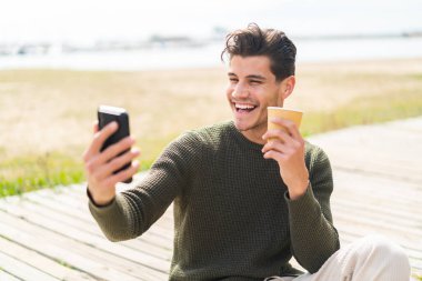 Açık havada cep telefonu kullanan ve elinde mutlu bir ifadeyle kahve tutan genç beyaz bir adam.